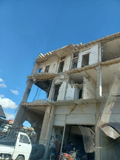 This image depicts a multi-story concrete structure with severe structural damage, likely located in Al-Qadam Municipality, Syria. The building, which appears to be at least four stories high, exhibits extensive crumbling concrete, exposed rebar, and missing wall sections across its upper floors, indicating significant damage from a collapse or destructive event.

On the ground floor, a commercial space operates beneath a relatively intact corrugated metal awning. A partially open metal roll-down door reveals an interior containing multiple circular metal objects mounted on a wall, alongside various tools and equipment, including what appears to be an engine or pump, and two large black tires stacked externally. Two individuals are present inside this ground-level space; one person, wearing dark clothing, is standing with their back to the viewer, while another is partially visible behind them.

To the left of the damaged building, a white utility pickup truck with a caged bed is partially visible. Further left, a small solar panel array is mounted on a rooftop. The scene is illuminated by a clear blue sky with sparse white clouds. The foreground on the right side of the image shows the interior of a vehicle, specifically a portion of the dashboard and windshield, indicating the perspective of the photographer.