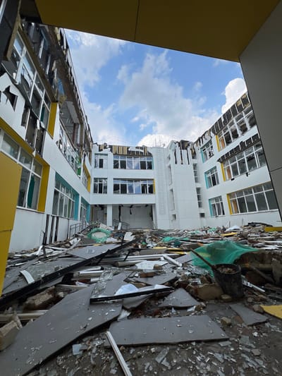 The image presents the severely damaged interior courtyard of a school building in Staryi Saltiv, Ukraine. The multi-story structure exhibits extensive destruction: its white facade, accented by yellow and light blue sections, is marred by missing and warped panels, exposing internal elements. Nearly all windows are shattered, with countless glass shards and frames scattered across the ground. The upper portions of the building show significant structural compromise and partial collapse. The entire courtyard floor is blanketed with a dense accumulation of varied debris, including large fragments of facade material (white, yellow, dark grey), shattered glass, concrete rubble, and twisted metal. Patches of green netting are visible amongst the refuse. In the foreground-right, a black bucket, possibly containing debris, rests beside a long-handled tool. The scene is observed under a partly cloudy daytime sky. No individuals are present.
