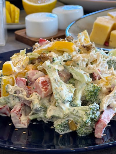 The image shows a close-up of a broccoli salad in a blue bowl. The salad also contains yellow peppers, red peppers, and nuts, all coated in a creamy white dressing. To the left of the bowl, partially visible, are cheese cubes and what appear to be sliced lemons. The location is not identifiable as Lermontovo, Armenia, based solely on the visual information in the image.