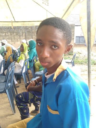 The scene is an outdoor gathering in Ijoko, Nigeria, taking place under a light-colored tent canopy. In the foreground, a young individual with dark skin and short, dark hair wears a blue collared shirt featuring a light brown trim on the collar and a breast pocket, looking directly into the camera with a neutral expression. Behind this individual and extending to the left, several people, primarily women wearing various colored hijabs—including light green, dark green, and yellow—and white or light-colored clothing, are seated in rows on blue plastic chairs. One woman in a light green hijab and white top is visible looking towards the camera. Adjacent to the foreground subject, the lower body of another individual is visible, clad in patterned blue and green trousers. A light-toned concrete block wall, bearing indistinct dark markings or text, forms part of the background structure beneath the tent. The ground surface is exposed earth. Partial text, including "OSORO" and a numerical sequence, is visible on a banner-like strip attached to the underside of the tent canopy.