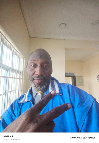 An adult male with dark skin, a bald head, and a graying beard is depicted in an indoor setting in Gwammaja, Nigeria. He is wearing a light gray collared shirt with a visible yellow top button, covered by a bright blue zip-up jacket featuring white and blue striped accents on the collar. His right hand is raised in the foreground, forming a "V" sign with his index and middle fingers, knuckles facing the camera. He looks directly forward. The background consists of cream-colored walls and ceiling. To the left, a barred window with white vertical and horizontal elements allows natural light. To the right, a partial view of an open doorway is visible, along with a rectangular, light-colored object, possibly a sign or document, mounted on the wall. A circular indentation and part of a white ceiling fan are discernible on the ceiling. Electrical conduits or cracks are visible at the wall-ceiling junction.