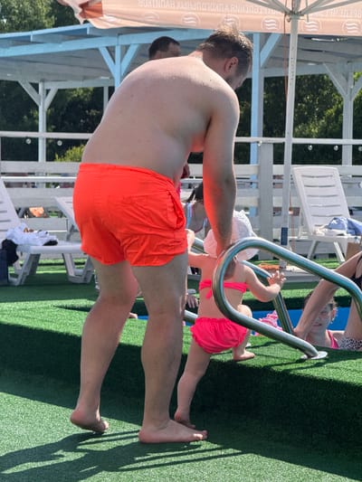 The image shows a family at a poolside in Koloksha, Russia. A man in orange swim trunks is in the foreground, his back to the camera. A young child in a pink bikini is climbing a pool ladder, being assisted by an adult whose arm is visible. Another child in a white hat and pink bikini bottom is near the ladder, and partially visible is another adult in a leopard print swimsuit. The setting appears to be a sunny day at an outdoor pool, with green artificial turf surrounding the water. White lounge chairs and a shaded area with a banner are visible in the background. The banner has text which appears to be in Cyrillic. The overall mood seems relaxed and family-oriented, enjoying a day at the pool.