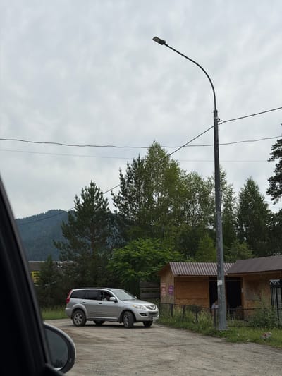 A silver SUV is parked on a dirt road next to a small wooden building with a corrugated metal roof. A person is visible inside the building. The scene is outdoors, with trees and a mountain in the background. A street lamp stands on the right side of the image. The sky is overcast. This appears to be the village of Ust-Sema, Russia, given the context.