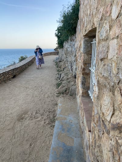 A person is standing on a path alongside the sea in Palafrugell, Spain. The person is wearing a hat, a blue floral dress, and a denim jacket. The path is bordered by a low stone wall on one side and a rough stone structure above. A glimpse of the blue sea and sky is visible. The scene is taken from a slightly elevated perspective, looking down the path.