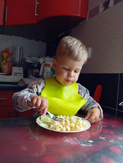 A young boy with blond hair is sitting at a table, eating pasta with a fork. He is wearing a yellow bib and a long-sleeved shirt with a cartoon pattern. The table has a red and black patterned surface. In the background, there are red kitchen cabinets, a blender, and various food items. The scene appears to be in a kitchen. The location is Hlynytsi, Ukraine.