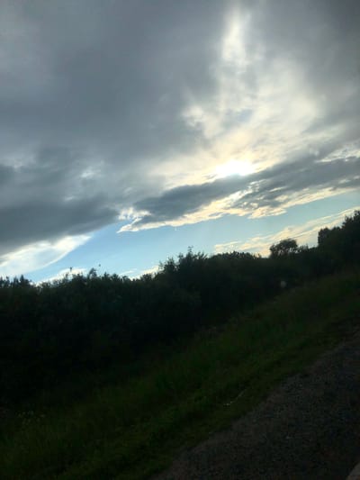 The image captures a natural landscape with a focus on the sky and a dense line of dark green foliage. The sky is partially obscured by dark, heavy clouds on the left, transitioning to lighter, thinner clouds with streaks of sunlight breaking through on the right. The sun is visible as a bright, hazy orb in the upper right quadrant, with rays of light radiating outwards. Below the sky, a continuous band of dark green vegetation, likely trees and bushes, stretches across the image. The foreground features an incline of darker, rough terrain, possibly gravel or dirt, with some patches of grass. The image appears to be taken outdoors, possibly in a rural or undeveloped area, and does not contain any people or specific urban structures. The overall mood is dramatic and somewhat overcast, with the sun attempting to penetrate the cloudy sky. Without further information, it is not possible to determine the specific location in Panino, Russia.