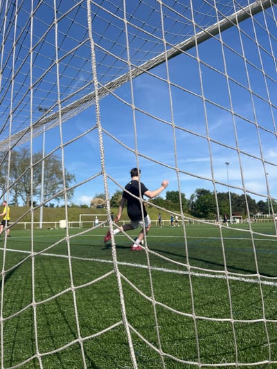 The image shows a soccer field in Champigny-sur-Marne, France, viewed through a soccer goal net. A male wearing a black shirt and light-colored shorts is in mid-air, possibly kicking a ball. The field is green with white lines. There are trees and other people visible in the background. The sky is blue.