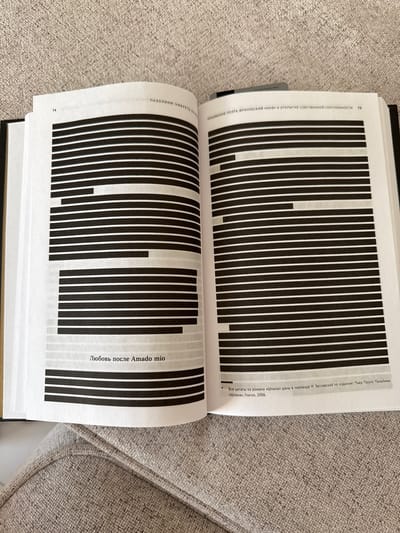 An open book rests on a grey, textured fabric surface, likely a sofa or chair. The book is oriented with its spine at the bottom, and the image is slightly rotated clockwise. Both visible pages are filled predominantly with black vertical bars of varying lengths, creating an abstract, barcode-like pattern, with white space in between and at the edges.
On the top left page, the page number "74" is visible, along with the partial text "ПАЗОЛИНИ. УМЕРЕТЬ ЗА" (PASOLINI. TO DIE FOR). The right page features a heading "ПРИЗВАНИЕ ПОЭТА, ФРИУЛЬСКИЙ «МИФ» И ОТКРЫТИЕ СОБСТВЕННОЙ СЕКСУАЛЬНОСТИ 75" (THE CALLING OF THE POET, THE FRIULIAN "MYTH" AND THE DISCOVERY OF ONE'S OWN SEXUALITY 75). Below the black bars on the right page, the text "Любовь после Amado mio" (Love after Amado mio) is visible. A footnote at the bottom right of the page reads: "Все цитаты из романа «Шпана» даны в переводе И. Заславской по изданию: Пьер Паоло Пазолини. «Шпана», Глагол, 2006." (All quotes from the novel "Ragazzi di vita" are given in I. Zaslavskaya's translation from the edition: Pier Paolo Pasolini. "Ragazzi di vita", Glagol, 2006.) The book appears to be a publication by or about Pier Paolo Pasolini, possibly with redacted or stylized text. The setting is indoors, with no specific time of day or weather discernible. The location context is Родниковское сельское поселение, Ukraine.