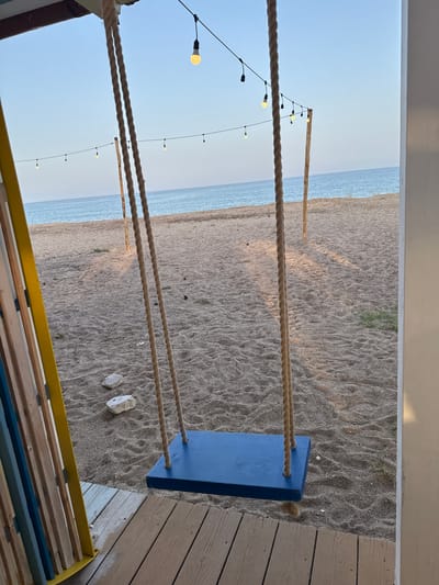 A swing with a blue seat hangs in the foreground, suspended by thick ropes from an unseen structure. The swing is positioned on a wooden deck, with the sandy beach and the sea visible beyond. The beach is scattered with small rocks. A string of lights with glowing bulbs stretches across the background, strung between two wooden posts. The sky is clear and blue. The scene appears to be located in Orosei, Italy, likely on the coast, given the beach and sea setting.