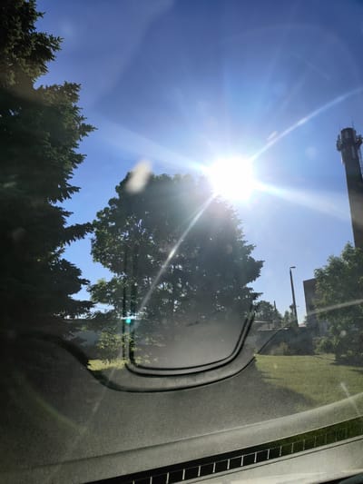 The image, captured from inside a vehicle looking out through the windshield, shows a bright sunny day. The sun is intensely bright, creating lens flare and glare across the image. On the left, a large evergreen tree dominates the foreground. In the center, another large deciduous tree is partially obscured by the sun's glare. To the right, a tall industrial tower, likely a communication or industrial chimney, is visible against the clear blue sky. A modern street lamp and some low buildings are also present in the background. The view suggests an outdoor setting, possibly in an industrial or suburban area of Saue, Estonia, given the presence of the tower and the general landscape. The lower portion of the image shows the interior of the car's dashboard and windshield, with reflections and glare. There are no people visible in the image.