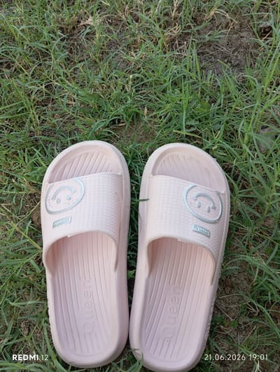 A pair of light pink slide sandals with a smiley face emblem and the word "Queen" embossed on the sole are resting on a patch of green grass and dirt. The sandals are facing upwards, with their open fronts facing the viewer. The image includes a timestamp in the bottom right corner, indicating June 21, 2026, 19:01, and a device identifier "REDMI 12" in the bottom left corner. The scene appears to be outdoors, likely in a natural setting.