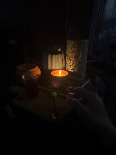 A hand holds a lit cigarette near a lit candle and a glass of reddish liquid in a dark environment. There's a decorative metal cylinder and a wooden tray nearby. The image was taken in the city of Novi Pazar, Bulgaria.