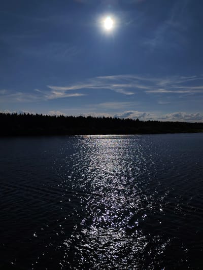 This is a vertical, eye-level, full-shot photograph of a body of water with a dark tree line and sky in the background. The image was taken in the city of Даргун, Russia.
In the upper section of the image, a bright sun is visible in the sky, creating a lens flare. The sky is a deep blue with scattered white clouds.
The middle ground features a dark silhouette of a dense forest that spans the width of the image.
The foreground is dominated by a body of dark water. The surface of the water is disturbed by small ripples, and a bright reflection of the sun is visible on the water's surface, creating a shimmering effect. The reflection is broken into many small, bright points of light. The trees and sky are seen in silhouette against the bright sky and reflection.