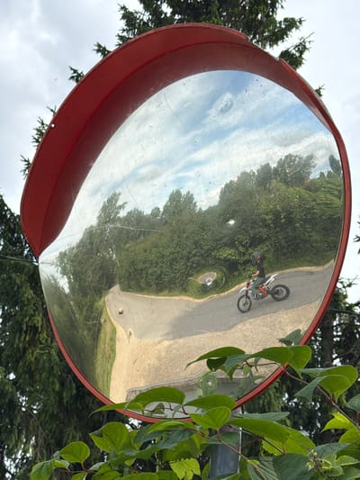The image shows a convex mirror mounted on a red frame, reflecting a rural road winding through a dense forest. A person on a white dirt bike, wearing a black shirt, turquoise pants, and a helmet, is riding on the road in the reflection. The sky above is overcast with scattered clouds. The mirror is surrounded by green foliage, and tall coniferous trees are visible behind it. The location is Tēraudi, Latvia.
