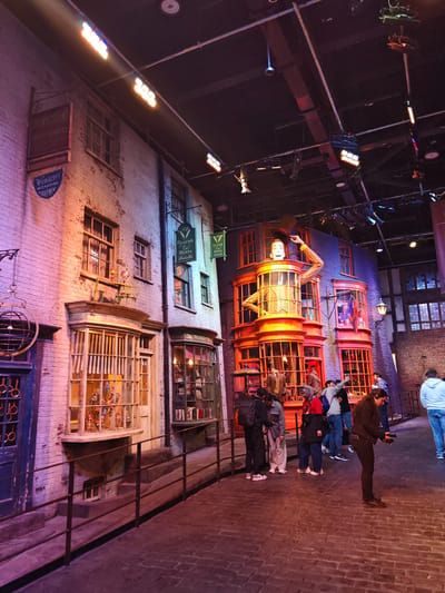 The image shows a street scene recreation of Diagon Alley at the Warner Bros. Studio Tour London in Watford, United Kingdom. On the left are the façades of shops with detailed windows. A black metal railing runs along a low ramp. People, including a woman holding a device, are looking at the attractions. The tall façade of Weasley's Wizard Wheezes is prominent in the center. Overhead, there are various stage lighting fixtures attached to the ceiling structure. The overall lighting creates a theatrical atmosphere.