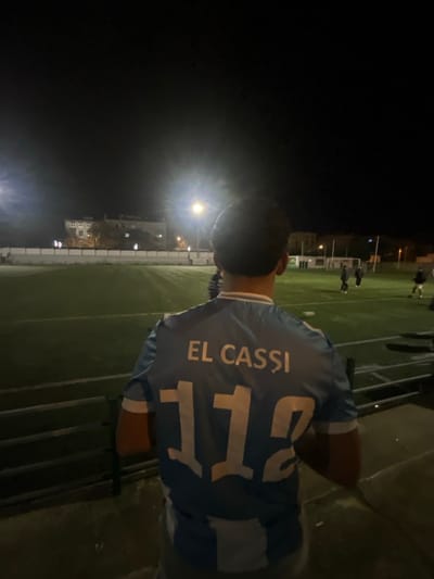 A blurry image shows the back of a person wearing a blue and white soccer jersey. The name "EL CASSI" is printed above the number "112" on the jersey. The individual stands near a soccer field at night, where a game appears to be in progress under stadium lights. The overall image has a low-light quality and a strong depth of field effect.