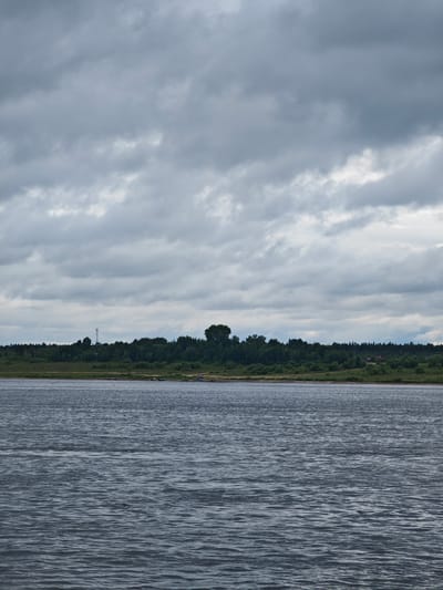 The image depicts a wide river flowing in Zotino, Russia, under a cloudy sky. The water is a dark, choppy gray, reflecting the overcast conditions. Along the far bank, a dense line of green trees stretches horizontally across the frame, with a few scattered buildings and a telecommunications tower visible amidst the foliage. The shoreline is a strip of light brown earth interspersed with green grass. Several small boats are moored or floating near the bank. The overall atmosphere is somber and natural, characteristic of a northern Russian landscape.