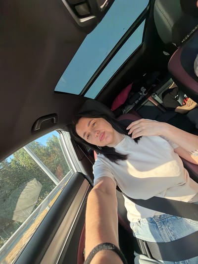 A woman is sitting in the passenger seat of a car, looking towards the camera. She has dark hair, fair skin with freckles, and is wearing a light-colored t-shirt and blue jeans. A seatbelt is fastened across her chest. The car has a panoramic sunroof, through which the sky and trees are visible. The interior of the car appears to be a dark color. The image appears to be taken during the daytime, with sunlight casting shadows. The location is identified as Uvarovo, Russia.