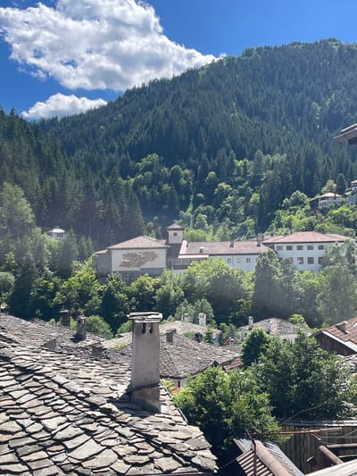 The image captures a picturesque view of Shiroka Laka, Bulgaria, showcasing traditional Rhodope architecture nestled within a verdant, mountainous landscape. In the foreground, weathered, slate-tiled roofs with prominent stone chimneys dominate the scene, indicating an older, well-established settlement. The roofs are layered and uneven, typical of historical building techniques.
Beyond the foreground, a cluster of larger buildings with red-tiled roofs emerges, interspersed with lush green trees. One prominent building features a white facade and a distinctive mural depicting a stylized, possibly mythological, figure or motif. Another building to its right is a larger, light-colored structure with multiple windows. These buildings appear to be part of a more institutional or public complex, perhaps a school or cultural center, given the mural and the scale.
The backdrop is a dense, rolling mountainside covered in a thick forest of evergreen trees, primarily pines. The forest stretches upwards, meeting a bright blue sky dotted with fluffy white clouds. The overall impression is one of tranquility and natural beauty, with the traditional village seamlessly integrated into the mountainous environment. The scene suggests a warm, sunny day, with the light casting shadows and highlighting the textures of the roofs and foliage. No people are visible in the image.