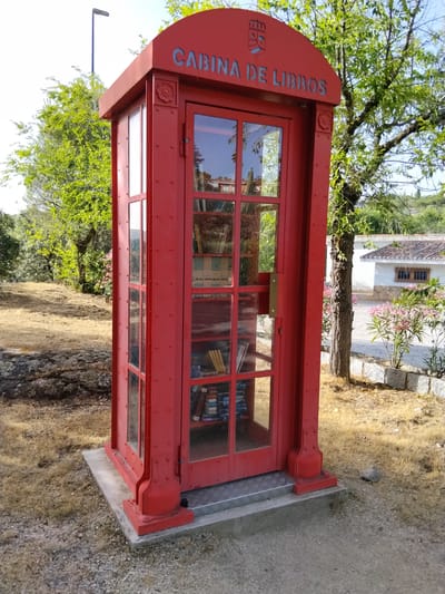 A bright red, glass-enclosed book exchange booth, resembling a classic telephone booth, stands on a concrete base in an outdoor setting. The booth is adorned with a coat of arms and text reading "CABINA DE LIBROS" in blue lettering at the top. Inside, shelves are filled with books, some visible through the glass panels. The booth is situated on a patch of dry, yellowish grass with scattered rocks and gravel. In the background, a white building with a tiled roof and wooden window frames is visible, surrounded by green trees and bushes. The scene appears to be in a park or public space in Hoyo de Manzanares, Spain. The lighting suggests it is daytime, with sunlight illuminating the area. There are no people or animals present in the frame.