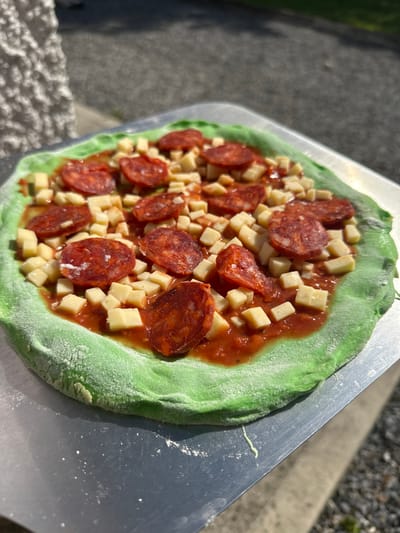 A close-up, overhead shot shows a green-crusted pizza topped with diced cheese, tomato sauce, and slices of pepperoni. The pizza rests on a metal peel, likely ready for baking. The background is blurred, suggesting an outdoor setting with sunlight dappling on a textured surface, possibly a sidewalk or stone wall. The specific location in Moortown, Ireland, is not discernible from the visual information provided.