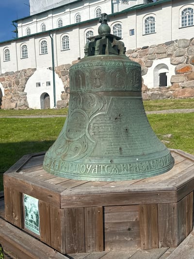 A large, ornate bronze bell sits on a wooden pedestal in front of a white stone building with arched windows. The bell is covered in intricate carvings and inscriptions. The building appears to be a historical or religious site, possibly a monastery given the location. The sky is blue and the ground is covered in green grass. The overall scene suggests a historical landmark in Solovetskiy, Russia.