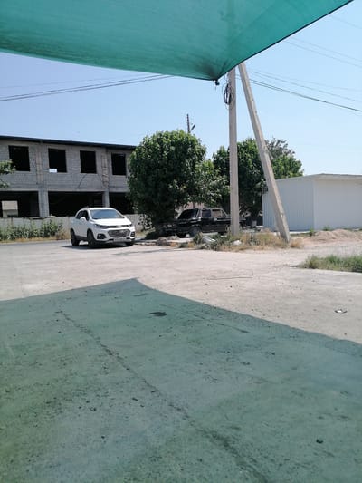 In the city of Gumbaz, Uzbekistan, a white Chevrolet Trax SUV is parked on a dirt lot, facing right, in front of a partially constructed building. The building, made of grey brick, has several open window frames. To the right of the SUV, a black sedan is parked. A large, white corrugated metal structure stands behind the sedan and to the right of the building. Green trees are scattered throughout the scene. Overhead, a green shade structure extends across the top of the frame, partially obscuring the clear blue sky. Electrical wires are visible, supported by a concrete utility pole. The ground is uneven and appears to be a mix of dirt and concrete.