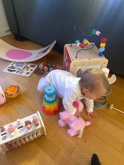 An infant, dressed in a white and light gray striped long-sleeved romper, is depicted in a crawling posture on a light-colored wooden floor within a residential setting in Rueil-Malmaison, France. The infant's head is turned to the right and slightly downward, with its right hand grasping a pink plush animal toy, which has a white tag attached.
The surrounding floor area is occupied by various children's toys. To the upper left, a curved light wood balance board, featuring a pink oval and a stylized animal face, rests against a dark gray upholstered sofa. Adjacent to the balance board is a flat wooden puzzle board with animal and plant-shaped cutouts. To the right of the puzzle, a segmented wooden caterpillar toy with rainbow-colored sections and wheels is positioned.
In the upper-right portion of the frame, a multi-sided wooden activity cube is visible, topped with a bead maze composed of blue wire tracks and various colorful wooden beads (red, yellow, green, pink, blue, orange). One visible side of the cube displays a rainbow illustration. Partially obscured to the right of the infant and near the activity cube are components of a light green and brown ball run or stacking toy, with a small wooden mallet lying beside it.
Directly to the infant's left is a conical stacking toy made of multi-colored, flower-shaped wooden pieces stacked in a rainbow sequence on a central peg. In the foreground, a white wooden hammering toy featuring several pink-tipped pegs and a set of pink-toned xylophone bars is present. Further to the left, a stack of colorful nesting bowls (yellow, orange, red, blue, green) is visible. The scene is illuminated by ambient light, suggesting an indoor daytime environment.