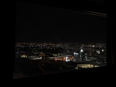 This is an aerial nighttime photograph of a cityscape, possibly from an elevated vantage point such as a high-rise building. The foreground is dominated by dark, silhouetted structures, suggesting a balcony or rooftop. Beyond this, a sprawling urban landscape unfolds, illuminated by a dense concentration of lights. Buildings of varying heights are visible, with some appearing to be modern high-rises and others lower-rise structures. The lights suggest residential areas, commercial districts, and possibly industrial zones. The overall impression is one of a busy, illuminated city at night, extending towards the horizon. The location is identified as Sao Thong Hin Town Municipality, Thailand, indicating a Southeast Asian urban environment.