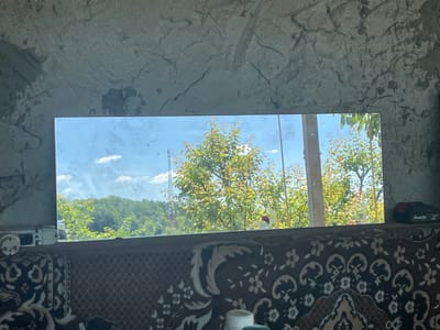 The image depicts a rectangular mirror set into a rough concrete wall. The mirror reflects a sunny sky with scattered clouds, and a landscape featuring lush green trees and foliage. In the background, a tall, slender radio tower is visible. The mirror is positioned above a patterned textile, possibly a rug or blanket, which covers a surface below. To the left of the mirror, a white electrical outlet or connector is attached to the concrete wall. To the right, a black and red power tool battery, with the brand name "Makita" visible, rests on the concrete surface. The scene appears to be an interior space with a view of an outdoor environment. The city of Velyka Berezovytsia, Ukraine, is mentioned in the prompt, but there are no explicit visual cues within the image to confirm this specific location.