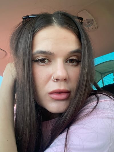A close-up, eye-level, selfie-style photograph shows a young woman with long, dark brown hair styled with a claw clip. She is wearing a pink shirt with a subtle pattern. The woman has brown eyes, winged eyeliner, and a silver nose ring. Her left arm is bent, with her hand resting near her head. The background suggests she is inside a vehicle, with part of the car's interior visible at the top, including a light fixture. The lighting is natural, casting a warm glow. The setting is Herasymivka, Ukraine.