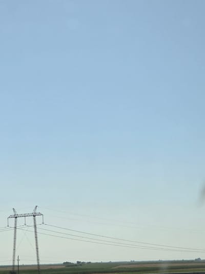 This image depicts a wide-angle view of a rural landscape under a clear, pale blue sky. In the foreground, a tall, lattice-style electricity pylon stands on the left side of the frame, supporting multiple power lines that stretch horizontally across the scene. The pylon is a metallic, dark structure with a complex, geometric design. Below the pylon, a smaller, dark pole is visible. The middle ground features agricultural fields, with visible rows of crops and patches of brown earth, suggesting a cultivated area. The horizon is low, with a faint line of trees and vegetation visible in the distance. The overall impression is one of open space and infrastructure in a rural setting, characteristic of the plains in the region of Jarak, Serbia.