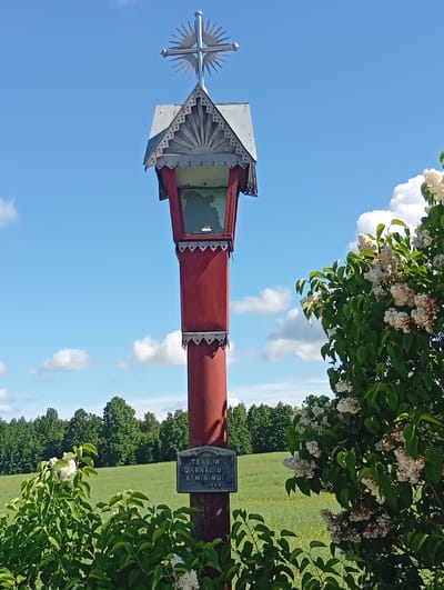 This is a low-angle, eye-level, medium shot of a red wooden wayside shrine with a metal cross on top, situated in Leičiai, Lithuania. The shrine is tall and slender, with decorative metalwork at the top and bottom of its red cylindrical body. Above the red section is a smaller, multi-faceted structure, also adorned with metalwork, topped by a star-shaped metal cross. A metal plaque is affixed to the shrine, near its base, bearing the inscription "TĖVŲ IR SENELIŲ ATMINTI" and "1999". The shrine stands in a grassy field, with lush green trees forming a dense line in the background under a bright blue sky dotted with white clouds. To the right of the shrine, a flowering bush with white and purple blossoms is prominent in the foreground, partially obscuring the shrine. To the left, another bush with white flowers and green leaves is visible. The overall impression is one of a peaceful, rural setting on a sunny day.