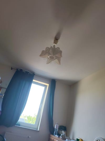 This is a photograph taken from a low angle, looking upwards into a room. The ceiling is white and illuminated by natural light coming from a large window. A decorative light fixture, possibly made of paper or a similar material, hangs from the ceiling. The fixture has a floral or petal-like design and is lit from within, casting a shadow on the ceiling.
To the left of the frame, dark green curtains are drawn partially across the window, which appears to be a casement window tilted open at the top. Through the window, trees and foliage are visible, suggesting an outdoor environment. Below the window, a white radiator is installed against the wall.
The right side of the image shows a corner of the room with a plain, light-colored wall. A wooden piece of furniture, possibly a dresser or cabinet, is visible in the lower right corner. On this furniture, there are several items, including a plastic bottle of water, a dark bag, a small white figurine resembling a rabbit, and some boxes. The room appears to be a residential interior, possibly a bedroom or living room, in Oława, Poland. The overall impression is one of a bright, domestic space.