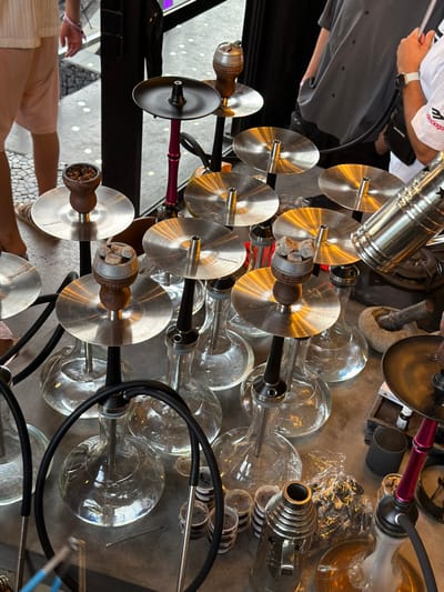 The image captures an elevated view of approximately ten shisha devices and related accessories arranged on a light-colored, reflective surface within an establishment in Tumbak Bayuh, Indonesia. Each shisha features a clear glass base, a black and silver stem, and a circular silver metallic ash tray, some bearing the "Hookah Tree" brand. Multiple ceramic bowls contain dark, granular material, presumed to be tobacco, with several topped by lit charcoal briquettes. Black rubber hoses, some with silver mouth tips, are coiled around the bases. To the right, a stone mortar and pestle set, several small plastic cups with dark contents, a metallic heat management device with star perforations, and a small wooden tray holding glass bottles are visible. Three individuals are partially depicted: one on the left in light shorts and sandals, another on the upper right wearing a grey button-up shirt, and a third on the far right in a white shirt with a "Balenciaga" logo and a white wristwatch. No direct interaction with the shishas by these individuals is discernible. The background suggests an interior space with a patterned floor.
