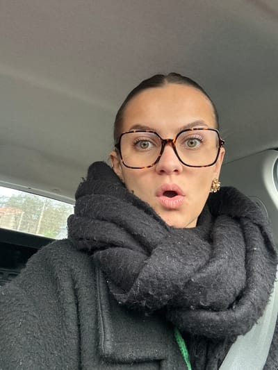 The image depicts a female individual inside a vehicle in the city of Saint-Cyprien, France. She is positioned centrally, viewed from the upper torso, with her head slightly angled. Her hair is dark and pulled back. She wears large, square-framed glasses with a tortoiseshell pattern, highlighting her light-colored eyes, which are wide open. Her mouth is agape in an "O" shape. A thick, dark, textured scarf is wrapped around her neck, partially obscuring a dark, textured coat. A gold-toned, clustered earring is visible on her right ear, and a small hoop earring on her left. A grey seatbelt diagonally crosses the lower right of the frame. The immediate background shows the light grey interior ceiling of the vehicle. Through the vehicle's left-side window, a blurred outdoor scene reveals muted green foliage and a light-colored building with a reddish roof under an overcast sky.