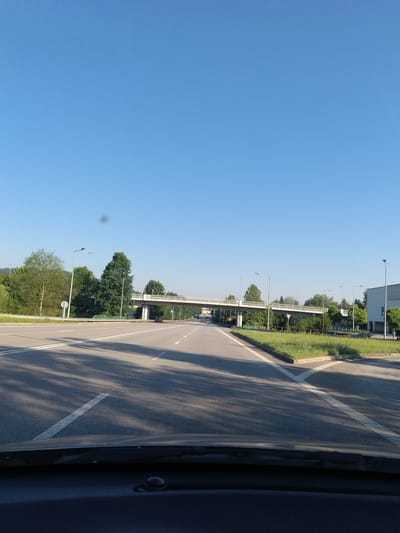 This media file depicts a view from a vehicle traveling along a highway on a clear, sunny day. The scene is characterized by a bright blue sky dominating the left side of the frame, transitioning into a grassy median and the road to the right. A white concrete overpass or bridge stretches across the scene in the middle ground, with trees and other vegetation lining the sides of the road and the area beneath the overpass. Several modern streetlights are visible, suggesting an urban or semi-urban environment. The road itself is asphalt with white lane markings. Shadows from unseen trees cast dappled patterns across the asphalt, indicating it is likely morning or late afternoon. No people are clearly visible. The overall impression is one of a typical daytime commute or travel through a well-maintained, modern road infrastructure area, possibly in a European setting given the location context. The image is oriented vertically, giving a sense of looking slightly downwards and sideways from the vehicle.