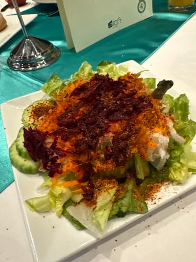 A square white plate holds a colorful salad. Green lettuce forms the base, topped with grated orange carrots, white shredded vegetables, and dark shredded beets. Round slices of cucumber ring the bottom. A reddish-brown spice is sprinkled generously over the salad. The plate sits on a table with a teal runner, next to a menu card with "Aloft" visible. Silver serving utensils are also present.