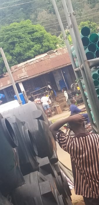 This image captures a bustling outdoor commercial area in Ado-Ekiti, Nigeria. In the foreground, multiple large, dark-grey plastic water storage tanks are stacked on the left, partially obscuring the view. To their right, the back of an individual wearing a brown and light-striped long-sleeved shirt is visible, with their right arm raised towards their head.

The midground features a single-story building with a rusty, corrugated metal roof and open-front structures, indicative of multiple small shops or market stalls. An individual in a white t-shirt and blue shorts stands near a small table displaying items for sale in front of this building. Another person in blue attire is partially visible to the right, possibly on a motorcycle or bicycle. Further back, a person in a white top and dark skirt, carrying a backpack, ascends steps leading to the building. Scattered blue plastic barrels and various goods are present near the structure.

On the far right of the midground, a rack holds numerous stacked green PVC pipes, suggesting a hardware or construction supply business. The background is characterized by a dense, green hillside covered in lush trees. Multiple utility poles with overhead wires extend vertically across the scene.