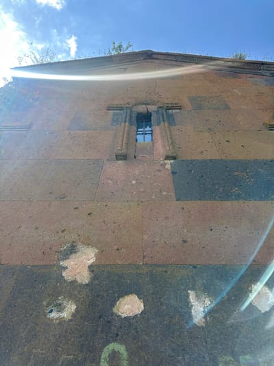 The image shows a close-up, low-angle view of the exterior wall of a stone building, likely a church, in Haykadzor, Armenia. The wall is constructed of large, rectangular stone blocks of varying reddish-brown and dark gray hues, laid in a somewhat irregular pattern. A small, arched window with dark panes is visible in the upper center of the frame, set within a decorative stone surround. The building exhibits signs of age and weathering, with some chipped stones and lighter patches on the surface, particularly in the lower section. The top of the wall shows an ornate stone frieze, and tufts of grass and weeds are growing along the roofline. The sky is bright blue with scattered white clouds. Sunlight creates a strong lens flare effect, casting bright streaks of light across the image. The overall impression is of an ancient, weathered structure set against a bright, sunny sky.