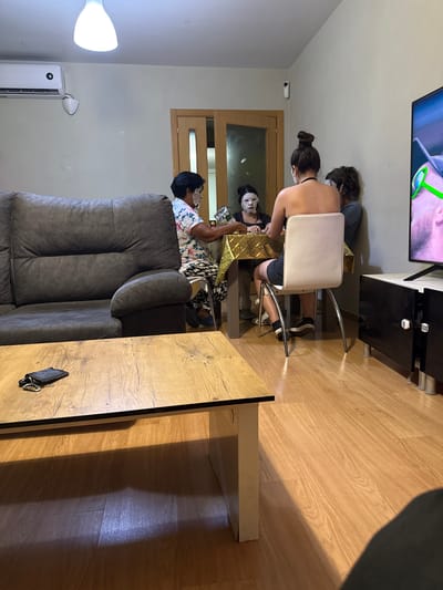 In a living room in Getafe, Spain, four individuals are gathered around a dining table, wearing face masks. Two of the individuals are women, and the other two are not clearly identifiable by gender. One woman, wearing a floral shirt and patterned leggings, is seated at the table, holding what appears to be a silver bag. Another woman, with her hair styled in a bun, is seated across from her, facing away from the camera. A third person, wearing a gray t-shirt, is seated next to the woman with the bun. The fourth individual, wearing a black shirt, is partially obscured by the table. The room features a dark gray sofa on the left, a wooden coffee table in the foreground with a black phone and keys on it, and a television on a black stand to the right. The floor is made of polished wood. An air conditioning unit is mounted high on the wall, and a light fixture hangs from the ceiling.