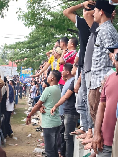 A medium shot captures a crowd of people from the waist down, standing and sitting on a concrete wall in Desa Meunasah Blang, Indonesia. The perspective is from the ground looking up, with the majority of the frame filled by people's legs and lower torsos. In the background, a banner with Indonesian text and images of people suggests a public event, possibly related to sports or a competition, as the text includes "SPORTIVITAS" (sportsmanship). The ground in front of the wall is littered with plastic bags, discarded food containers, and various pairs of footwear, indicating a casual outdoor gathering. The atmosphere appears to be one of observation, with people looking towards something out of frame. The overall scene is typical of a community event in Indonesia.