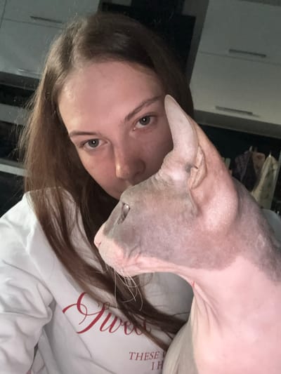 A young woman with long brown hair is captured in a selfie with a Sphynx cat. The woman is wearing a white sweatshirt with red lettering. The cat, with its characteristic wrinkled pink skin, is nestled close to the woman's face, its head turned to the side. The background appears to be an indoor setting, possibly a kitchen, with cabinets visible. The lighting is dim, suggesting it might be evening or a poorly lit room. The image's composition is intimate, focusing on the bond between the woman and her pet. No specific landmarks or details indicative of Novaya Ivanovka, Russia, are present in the image.