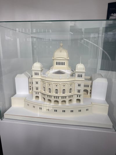 This media file shows a model of the Swiss Parliament Building in Bern, Switzerland. The model is detailed and appears to be made of a cream-colored material. It features a large central dome with a spire, smaller domes, multiple levels of windows, and arched walkways. The model is displayed inside a glass case, with reflections visible on the glass.
Behind the model, there is a black and white photograph with some text. The visible text indicates that the photo is related to "Bern boomt" (Bern is booming) and mentions "Das Parlament" (The Parliament) and "Baustelle von Westen" (Construction site from the West). Other text fragments suggest details about the building's construction period ("erbaut 1894-1902") and its location ("Standort am neu guschaffenen Bundesplatz" - Location on the newly created Federal Square).
The overall scene is that of an exhibit, likely in a museum or information center, showcasing the historical and architectural significance of the Swiss Parliament Building. The lighting is neutral, suggesting an indoor setting, possibly during the daytime. There are no people clearly visible in the main frame of the image, but the context of a historical exhibit implies a passive audience viewing the display. The model itself is the primary subject, highlighting the building's grand design.