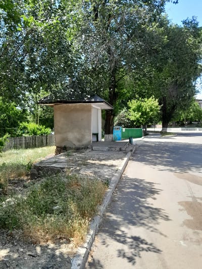 A bus stop shelter made of concrete and stucco stands on a concrete platform next to an asphalt road. The shelter has a dark, sloped roof and an open front with a green bench visible inside. To the left of the shelter, a weathered wooden fence runs along the edge of a grassy area. Beyond the fence, lush green trees and foliage create a dense backdrop. In the distance, a bright green metal gate is visible. The road is partially shaded by the dappled sunlight filtering through the leaves of overhead trees. The scene appears to be a quiet, somewhat rural or suburban setting in Cimișlia, Moldova.