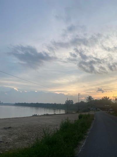 The image depicts a scenic view of a road alongside a body of water, possibly a river or lake, in Lembah Dusun Gading, Indonesia. The scene is bathed in the soft, warm light of either sunrise or sunset, indicated by the golden hues in the sky and the elongated shadows. Wispy clouds stretch across the sky, adding texture and depth to the atmosphere.

On the right side of the frame, an asphalt road runs alongside a strip of green grass. Beyond the grass, the water stretches out, reflecting the muted colors of the sky. The left bank of the water is lined with lush green vegetation and trees, creating a natural boundary. In the distance, a faint silhouette of mountains can be seen through a slight haze, suggesting a tranquil, natural setting.

Several utility poles with power lines are visible along the roadside and extending across the landscape, hinting at human presence and infrastructure in this otherwise natural environment. The overall mood of the image is peaceful and serene, capturing the quiet beauty of the Indonesian landscape at a transitional time of day. No people are clearly discernible in the image, but a couple of figures appear to be near the water's edge on the sandy bank. The image is oriented vertically, so the scene is viewed as if the camera was tilted.