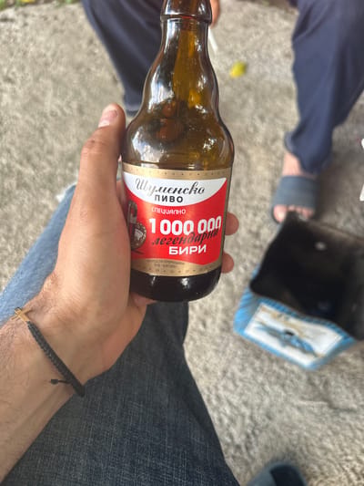 A man's hand, with a woven bracelet with a golden cross, holds a glass bottle of Shumensko Pivo beer. The label on the bottle is red and white with Cyrillic text that reads "Шуменско ПИВО СПЕЦИАЛНО 1000 000 легендарни бири". The background shows a carpeted floor, the legs of two people in blue pants, and a black bag with blue trim. The scene appears to be outdoors, possibly in Zimnitsa, Bulgaria.