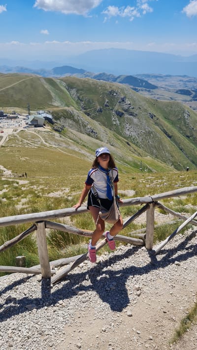 A young girl, wearing a blue baseball cap, a white and blue short-sleeved shirt with red trim, black shorts, and pink shoes, is perched on a wooden railing on a mountainside. She is looking up and to her right, with a light-colored crossbody bag slung over her shoulder. Her legs are bent, with her pink-shoed feet dangling over the railing. The railing is made of rough-hewn wooden posts and rails, with a rocky, gravelly path beneath it. The background reveals a vast mountainous landscape with rolling green hills, dotted with sparse vegetation and rocky outcrops. In the distance, several layers of hazy blue mountains fade into the horizon under a bright blue sky with scattered white clouds. To the left, a modern building complex with a white and gray structure and a parking lot is visible on a lower slope, suggesting it might be a ski resort or a visitor center. The scene appears to be located in the mountainous region surrounding L'Aquila, Italy, possibly at a high-altitude viewpoint.