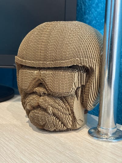 A layered cardboard sculpture of a human head rests on a metal railing. The background shows a light wooden panel and a dark-colored structure with blue accents. The location, Chuprino, Russia, is not evident from the image.