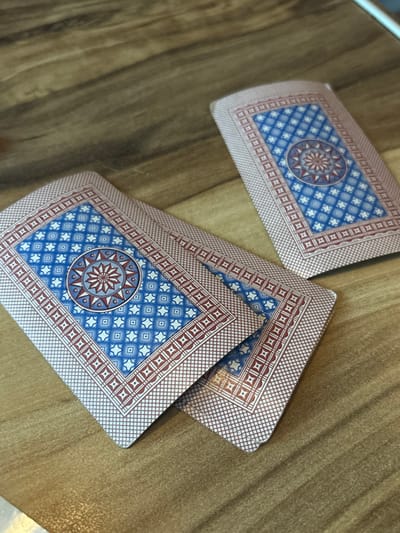 Three distinct groupings of playing cards are displayed face-down on a light-brown wooden surface in Pervomaiskoe, Russia. Two stacks of cards are positioned in the foreground, with one partially overlapping the other towards the lower-left of the frame. A third, smaller stack or single card is visible further back, towards the upper-right. All visible card backs feature an identical design: a central circular medallion with a multi-pointed star motif rendered in red, grey, and light blue, set against a dark blue background. This central element is encompassed by a field of light blue diamond patterns on a dark blue background, bordered by an inner strip of repeating red squares and an outer fine cross-hatched red border. The cards show signs of wear along their edges and corners, and a noticeable brown discoloration is present on the bottom right corner of the foremost foreground card. The wooden surface exhibits a distinct grain pattern, running predominantly horizontally across the frame. No individuals or active interactions are visible.